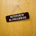 A képen az "Interview in progress", azaz "interjú folyamatban" tábla látható egy faajtóra vagy burkolati elemre akasztva.