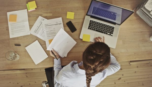 A képen egy hölgy látható, amint egy asztalnál ül, papírral a kezében és a laptopján dolgozik