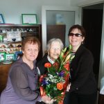 A képen középen a 104 éves ünnepelt, Margitka néni látható virággal a kezében. Mellette jobbról Fodor Ágnes elnök, balról Vasné Pintér Teréz közösségi civilszervező.