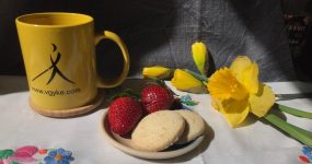 A képen: hímzett terítőn VGYKE logós sárga bögre, tálkában eper és vajas keksz, a háttérben bimbós nárciszok.
