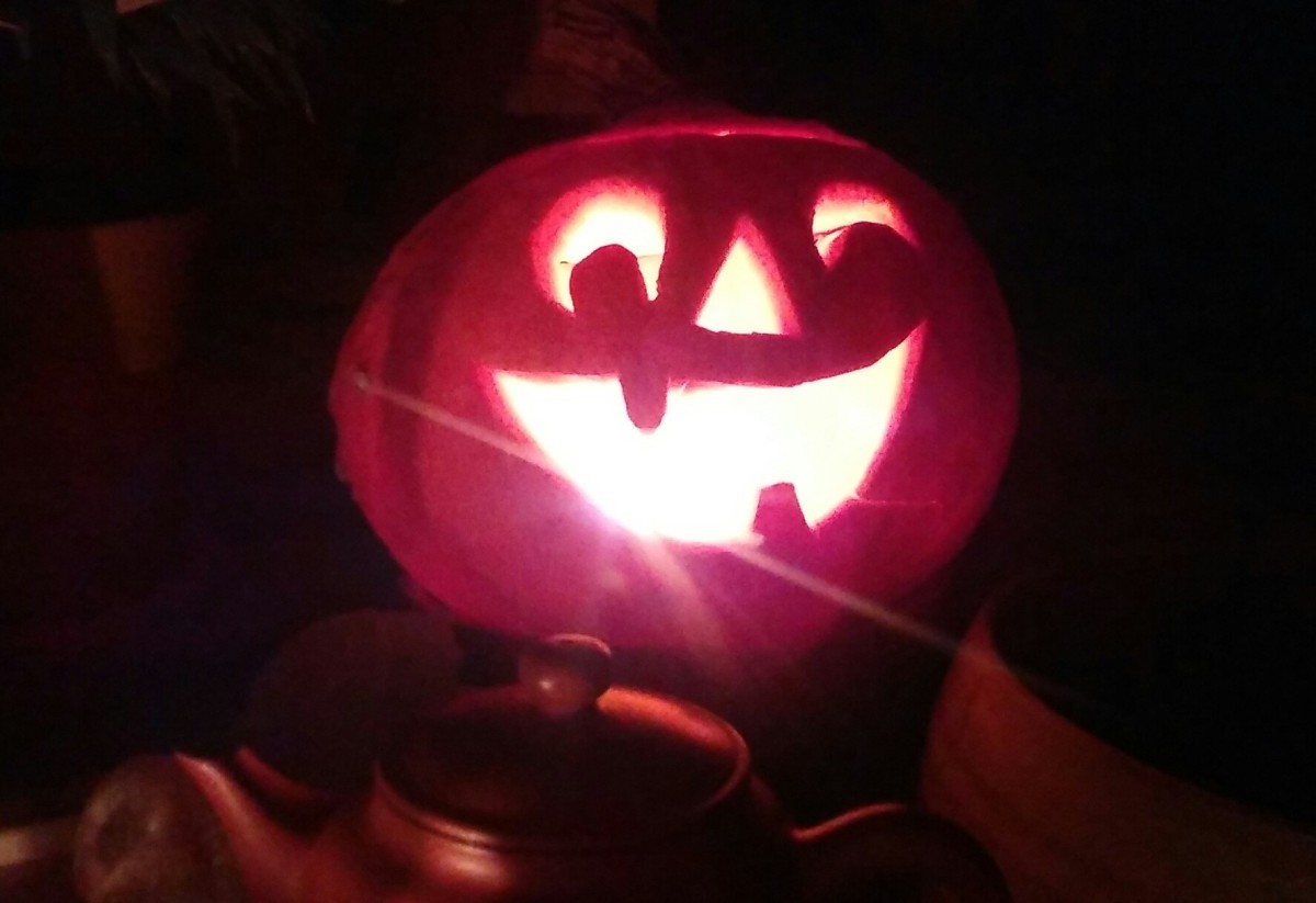 Őszbúcsúztató utó-utó-Halloween-buli jelmezversennyel - egy vigyorgó töklámpás a sötétben, előtte teáskanna teteje látszik.