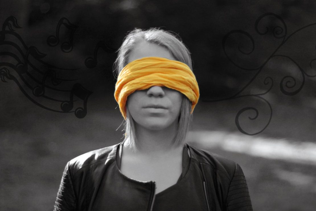 A képen egy lány látható fekete fehérben, egy sárga kendővel van bekötve a szeme.