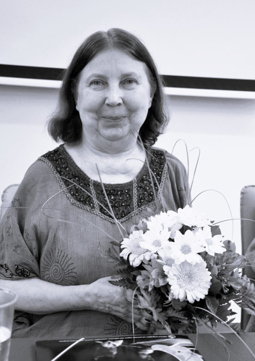 Elhunyt Dr. Gyimesiné Dobos Marianna - a kép fekete-fehér. Mariann mosolyogva egy virágcsokrot tart a kezében.