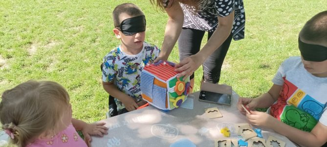Júniusi szemléletformáló programok - a gyerekek szemtakarót viselve játszanak az asztalnál. A program szabadtéri.